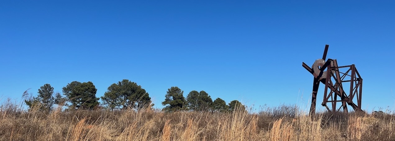 A photograph of a meadow under a cloudless blue sky. Some trees are in the distance. Closer and to one side is a curious metal sculpture, resembling a set of framework made of tumbled-together metal beams.